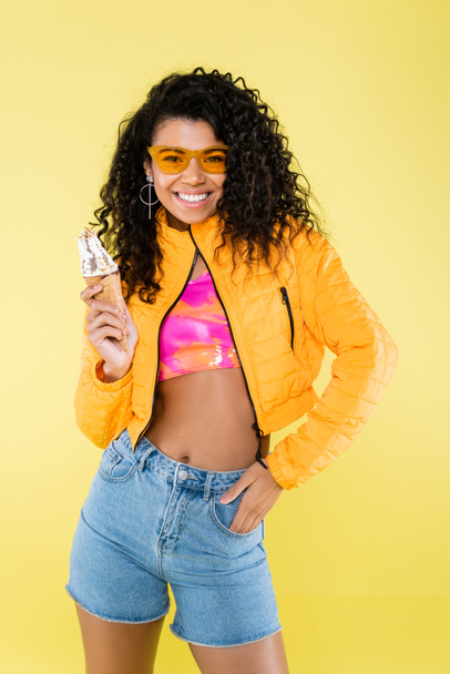happy african american young woman in sunglasses holding ice cream cone isolated on yellow - Valokuva, kuva