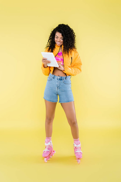 full length of positive african american young woman on roller skates using digital tablet on yellow - Photo, Image