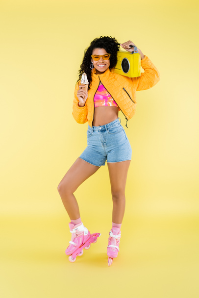 happy african american young woman in roller skates holding ice cream cone and boombox isolated on yellow - Fotoğraf, Görsel