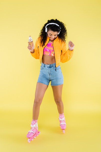 happy african american young woman in headphones and roller skates holding ice cream cone on yellow - Foto, Bild