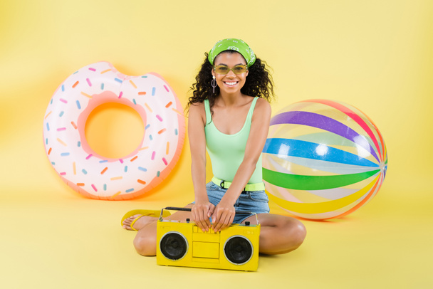 full length of happy african american woman sitting with boombox near inflatable ball and ring on yellow  - Photo, Image