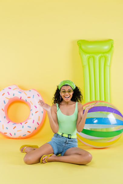 full length of cheerful african american woman sitting near inflatable ball, mattress and ring on yellow  - Fotografie, Obrázek