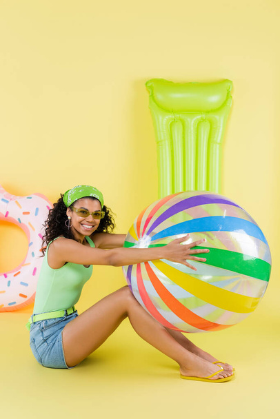 full length of joyful african american woman sitting with inflatable ball, mattress and ring on yellow  - Photo, Image