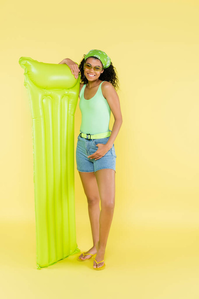 黄色の上に膨脹可能なマットレスを持つ陽気なアフリカ系アメリカ人の若い女性の完全な長さ  - 写真・画像