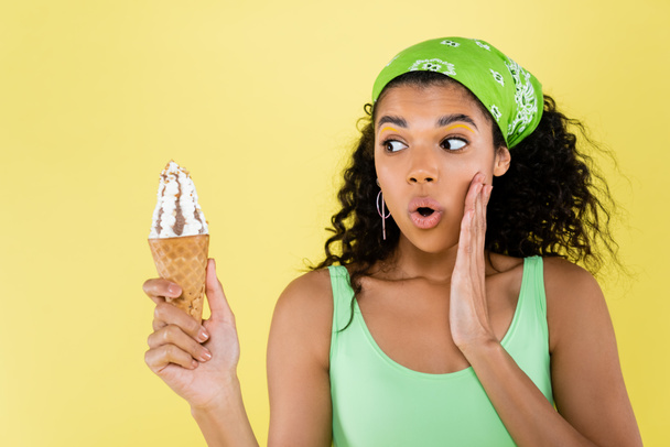 surprised african american woman in green kerchief looking at ice cream isolated on yellow - Φωτογραφία, εικόνα