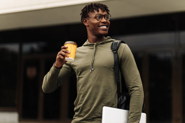 Stylish smiling African American man in glasses, drinking coffee in a cup and walking up the stairs in the city center with a laptop in his hands, stylish freelance people, with a laptop - Foto, imagen