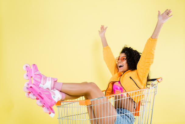 amazed african american young woman in pink roller skates sitting in shopping cart on yellow - Fotó, kép