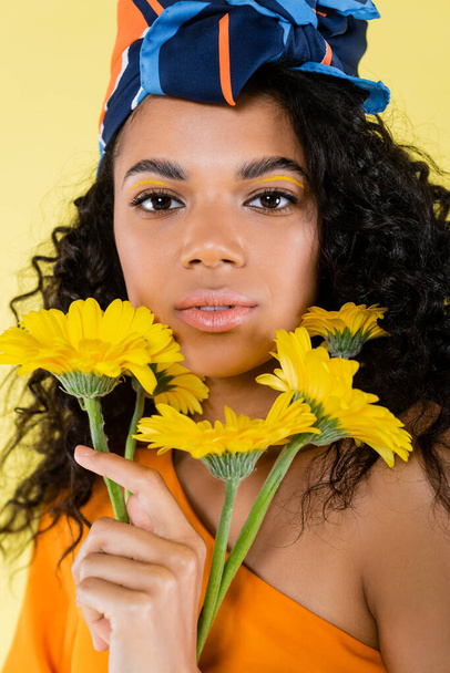 mujer joven afroamericana sosteniendo flores aisladas en amarillo - Foto, imagen