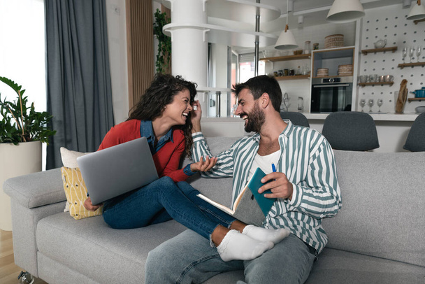 Loving young happy couple using laptop and analyzing their finances, writing notes, man with notebook woman with computer and having fun in their cozy home. Married people working from home. - Foto, immagini