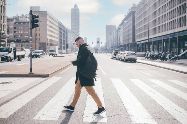 Young bearded caucasian man walking outdoor city crosswalk using smartphone wearing backpack, traveller checking map or commuter going work reading emails - Фото, изображение