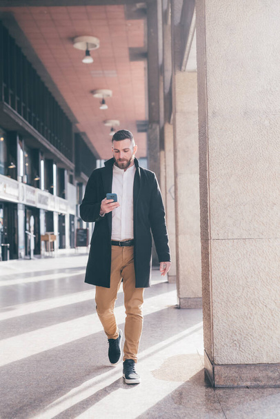 Νεαρός γενειοφόρος άνδρας που περπατά σε εξωτερικούς χώρους χρησιμοποιώντας smartphone βλέποντας βίντεο που ζουν streaming ή ψώνια online πελάτη ecommerce - Φωτογραφία, εικόνα