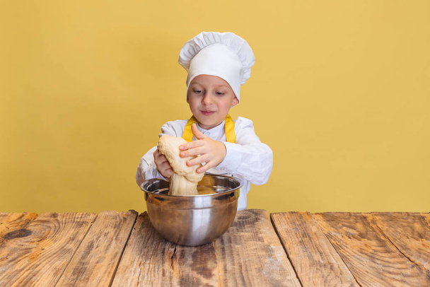 Cute smiling boy in white cook, chef jacket and hat kneads dough isolated on yellow studio background. - Φωτογραφία, εικόνα