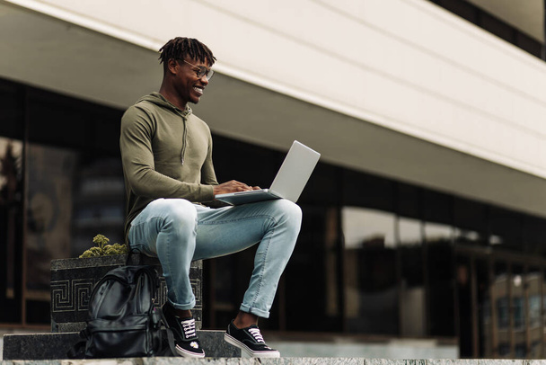 African black man working on a city street on a bench, uses a laptop computer for work, against the background of a modern building, office worker, freelancer. - Photo, Image