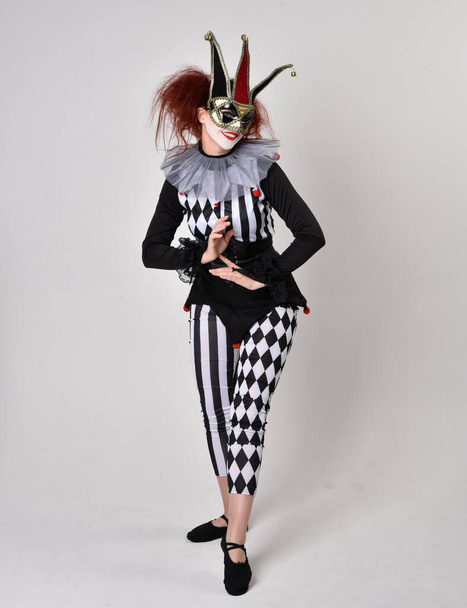 Full length  portrait of red haired  girl wearing a black and white clown jester costume, theatrical circus character.  Standing pose  isolated on  studio background. - Photo, Image