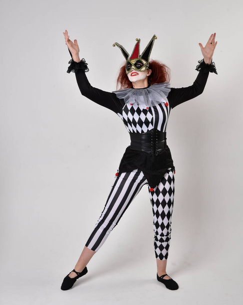 Полнометражный портрет рыжеволосой девушки в чёрно-белом костюме шута-клоуна, театрального циркового персонажа. Стоячая поза изолирована на фоне студии. - Фото, изображение