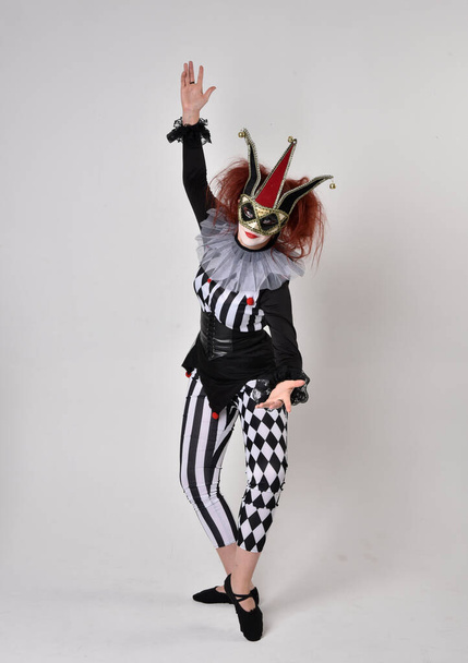 Retrato de cuerpo entero de una chica pelirroja vestida con un traje de bufón de payaso blanco y negro, personaje teatral de circo. pose de pie aislado en fondo de estudio. - Foto, Imagen