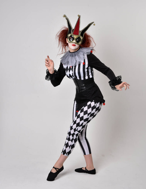 Полнометражный портрет рыжеволосой девушки в чёрно-белом костюме шута-клоуна, театрального циркового персонажа. Стоячая поза изолирована на фоне студии. - Фото, изображение