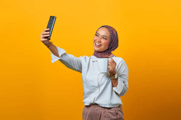 Alegre mujer asiática utilizando el teléfono celular haciendo selfie mientras sostiene taza sobre fondo amarillo - Foto, Imagen