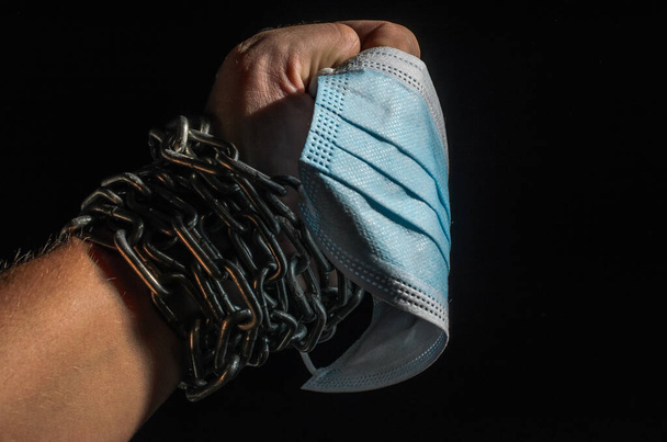 Human hand chained in chains holds a medical protective mask, isolated on black background - Foto, imagen