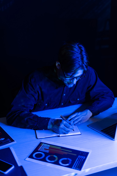 黒で区切られたテーブルの上の紙やデバイスの近くのノートブックに書く眼鏡のプログラマー  - 写真・画像