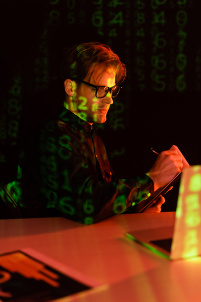 Programmer in eyeglasses writing on notebook near blurred laptop and reflection of computer code on black background  - Foto, afbeelding