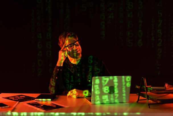 Uśmiechnięty programista w okularach patrzący na laptopa przy odbiciu kodu komputera na czarnym tle  - Zdjęcie, obraz