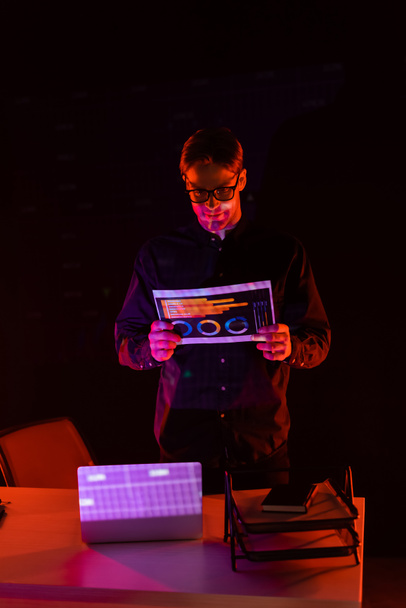 Młody programista trzymający papier z wykresami przy laptopie na stole odizolowany na czarno  - Zdjęcie, obraz