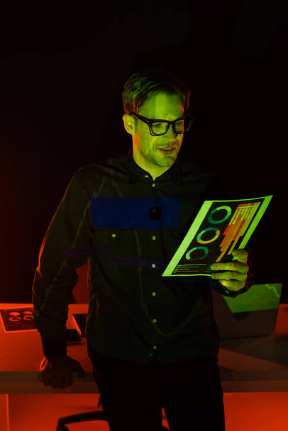 Programador sonriente sosteniendo documento con gráficos cerca del ordenador portátil con reflexión sobre fondo negro  - Foto, imagen