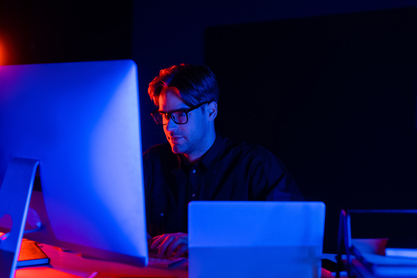 Joven programador en anteojos usando computadora cerca de laptop sobre fondo negro con iluminación  - Foto, imagen