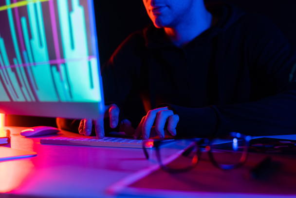 Cropped view of programmer using computer with charts reflection near eyeglasses on table on black background - 写真・画像
