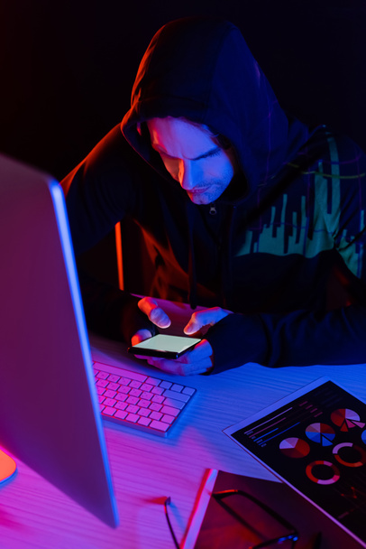 Hacker με αντανάκλαση των διαγραμμάτων χρησιμοποιώντας smartphone κοντά στον υπολογιστή και χαρτιά που απομονώνονται σε μαύρο  - Φωτογραφία, εικόνα