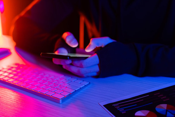 Cropped view of programmer using smartphone near computer keyboard and document on black background  - Photo, Image