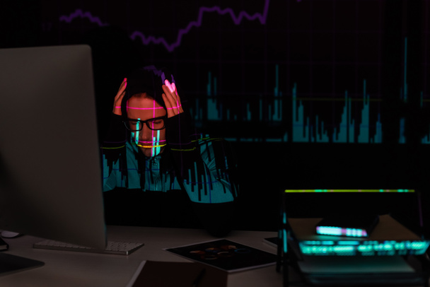 Upset hacker with charts reflection sitting near computer and documents on black background  - Foto, Bild