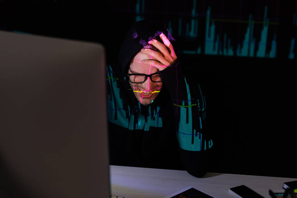 Θλιβερός χάκερ με αντανάκλαση των διαγραμμάτων κάθεται κοντά σε συσκευές σε μαύρο φόντο  - Φωτογραφία, εικόνα