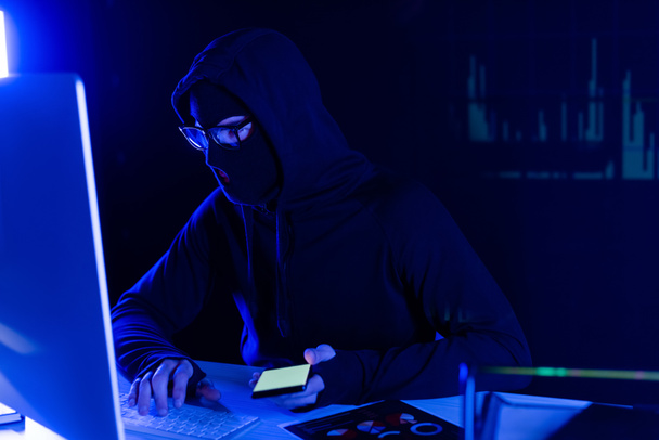 Хакер в балаклаве с помощью компьютера и смартфона на черном фоне с отражением диаграмм  - Фото, изображение