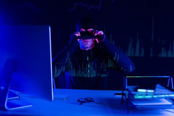 Haker nosi kominiarkę w pobliżu smartfona i komputera na czarnym tle z odbiciem wykresów  - Zdjęcie, obraz