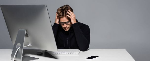 Programmatore scontento negli occhiali seduto vicino al computer e smartphone isolato su grigio, banner  - Foto, immagini