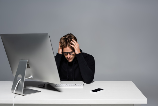 Расстроенный программист, сидящий рядом с компьютером и сотовым телефоном на столе изолирован на сером  - Фото, изображение