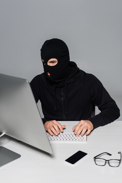 Hacker in balaclava using computer near smartphone and eyeglasses isolated on grey  - Photo, Image