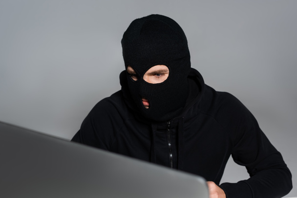 Haker w kominiarce patrzy na monitor komputera odizolowany na szaro  - Zdjęcie, obraz