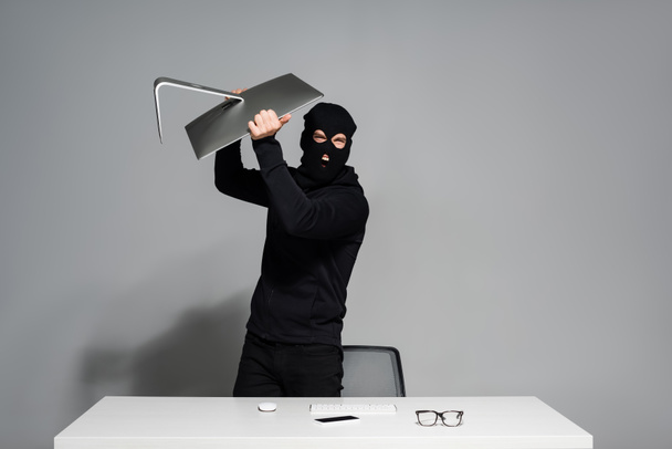 Angry hacker holding computer monitor near smartphone and eyeglasses on table on grey background  - Photo, Image