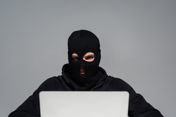 Hacker v balaklava při pohledu na notebook izolované na šedé  - Fotografie, Obrázek