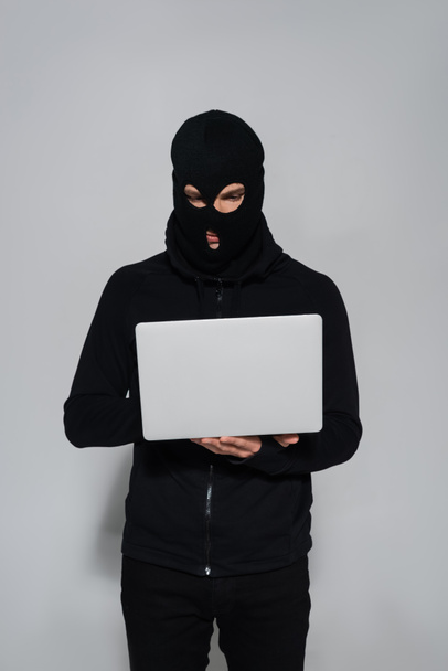Хакер в черной балаклаве с помощью ноутбука на сером фоне  - Фото, изображение