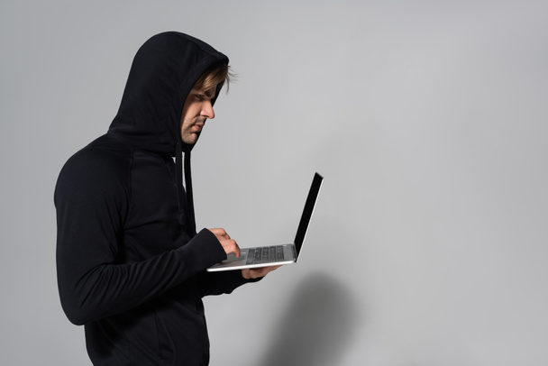 Seitenansicht des Hackers mit Laptop auf grauem Hintergrund  - Foto, Bild