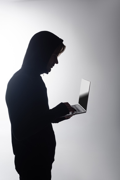 Boční pohled hackera v kapuci pomocí notebooku izolovaného na šedé  - Fotografie, Obrázek