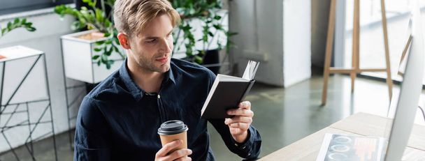 Young programmer looking at notebook while holding coffee to go near computer in office, banner  - Foto, Bild