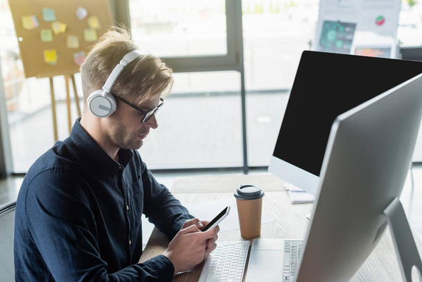 Vista lateral do desenvolvedor em fones de ouvido usando smartphone perto de café e computadores no escritório  - Foto, Imagem