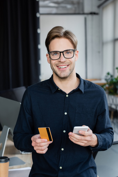 Glimlachende ontwikkelaar met creditcard en mobiele telefoon terwijl hij naar de camera op kantoor kijkt  - Foto, afbeelding