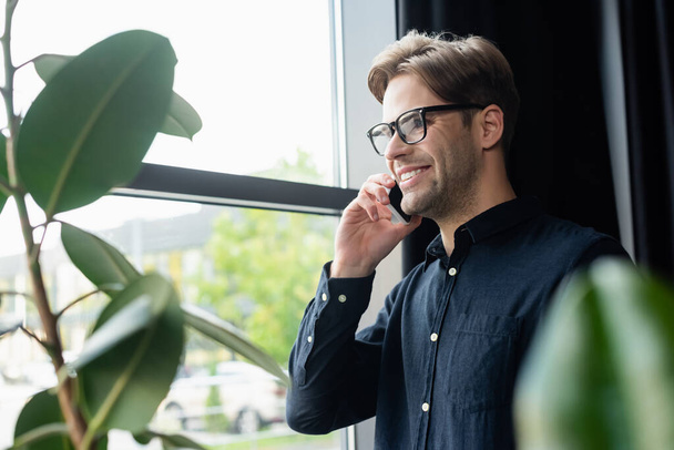 Positive programmer in eyeglasses talking on smartphone near plants in office  - Fotografie, Obrázek