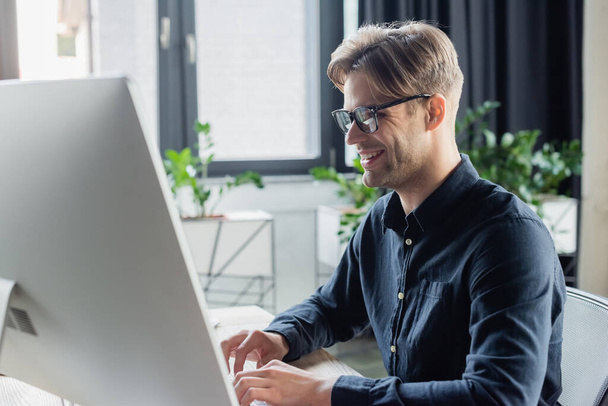 Smiling developer in eyeglasses using computer keyboard near monitor in office  - Fotó, kép